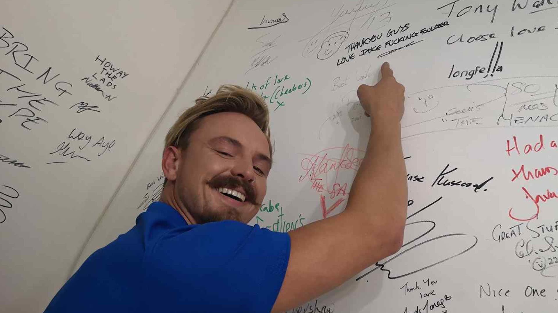 Man smiling and pointing at signatures wall.